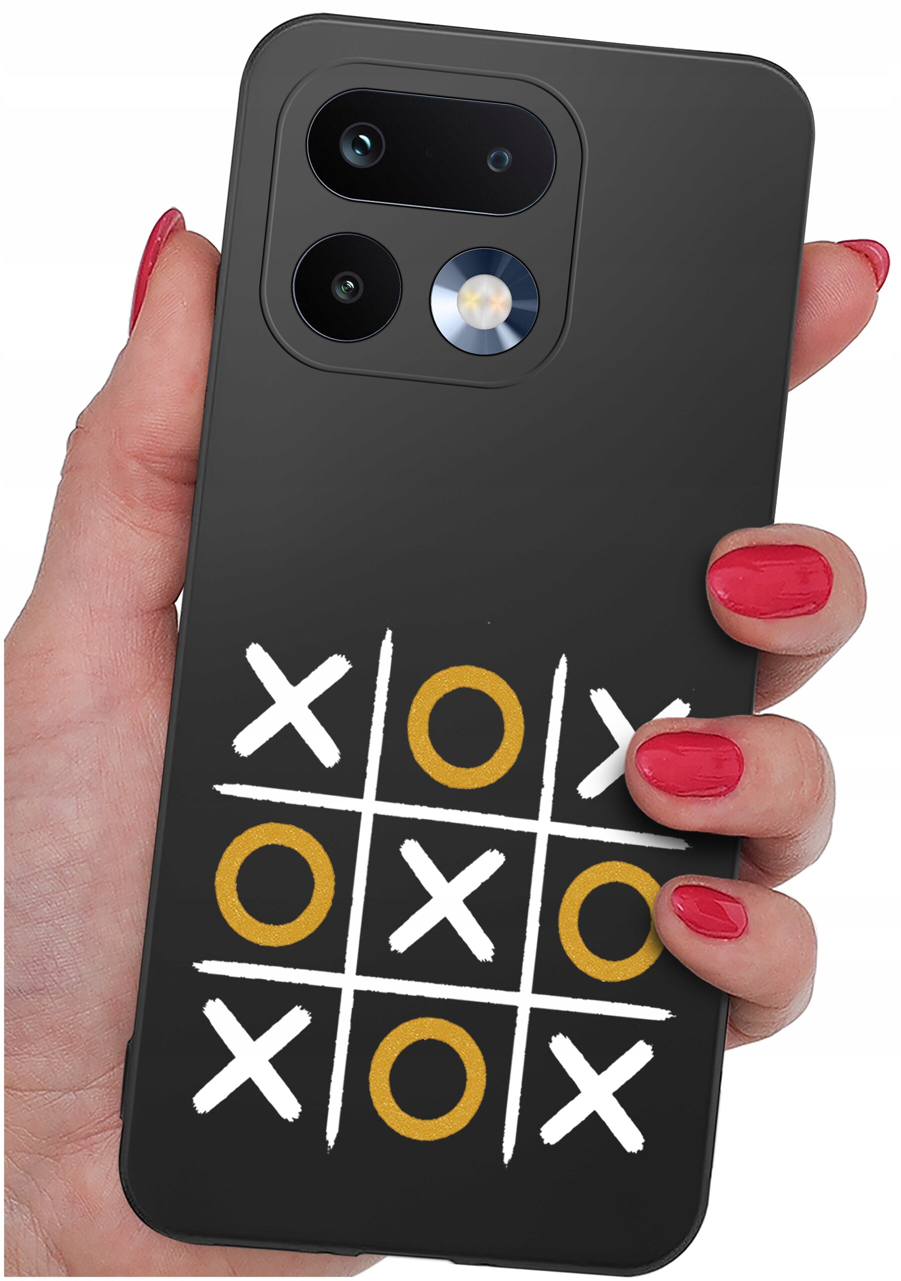 Czarna obudowa telefonu z nadrukiem gry w kółko i krzyżyk, trzymana w dłoni z czerwonymi paznokciami. Na obudowie widoczne są białe krzyżyki i złote kółka.