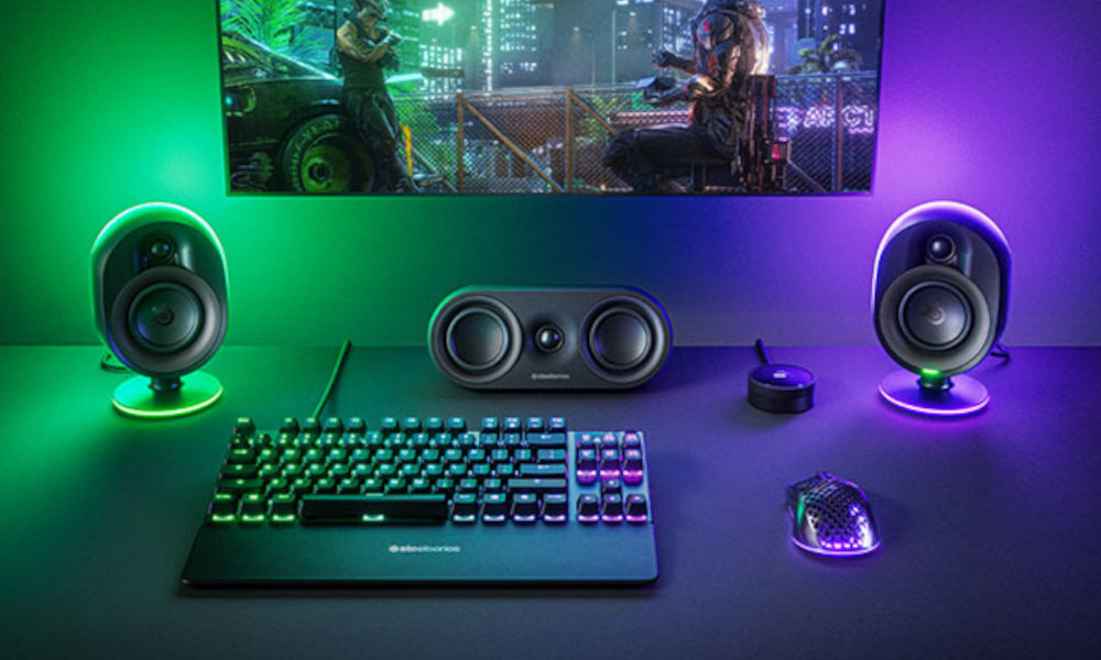 Na biurku znajdują się dwa głośniki z podświetleniem, klawiatura, myszka oraz centralny głośnik z logo SteelSeries. W tle widoczny jest ekran z grą komputerową.