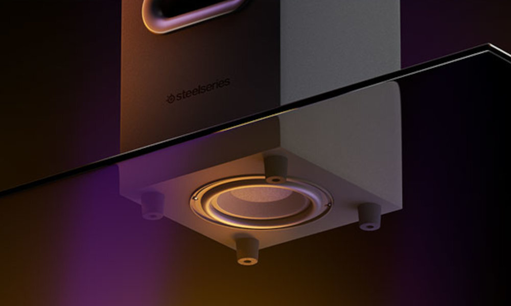 Obraz przedstawia głośnik z widocznym logo \'steelseries\' na obudowie. Głośnik jest umieszczony na szklanej powierzchni, a jego dolna część jest podświetlona ciepłym światłem.