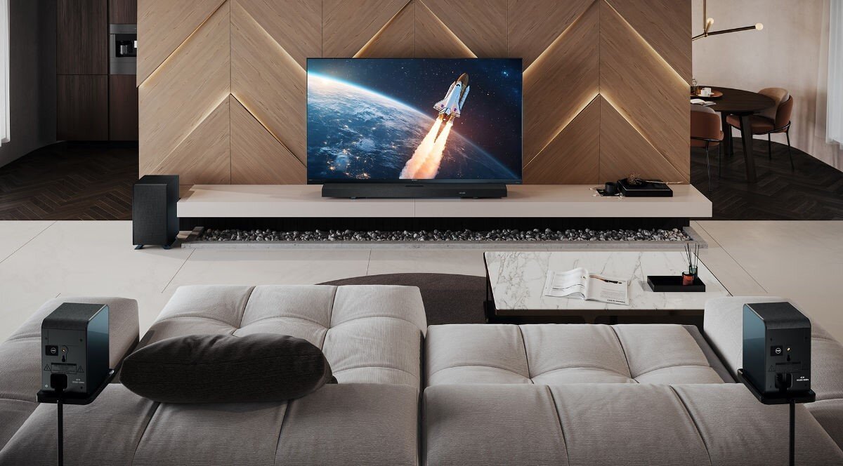 W salonie znajduje się duży telewizor wyświetlający obraz startującej rakiety, ustawiony na niskiej szafce. Przed telewizorem stoi jasna sofa, a po bokach widoczne są głośniki systemu dźwiękowego.