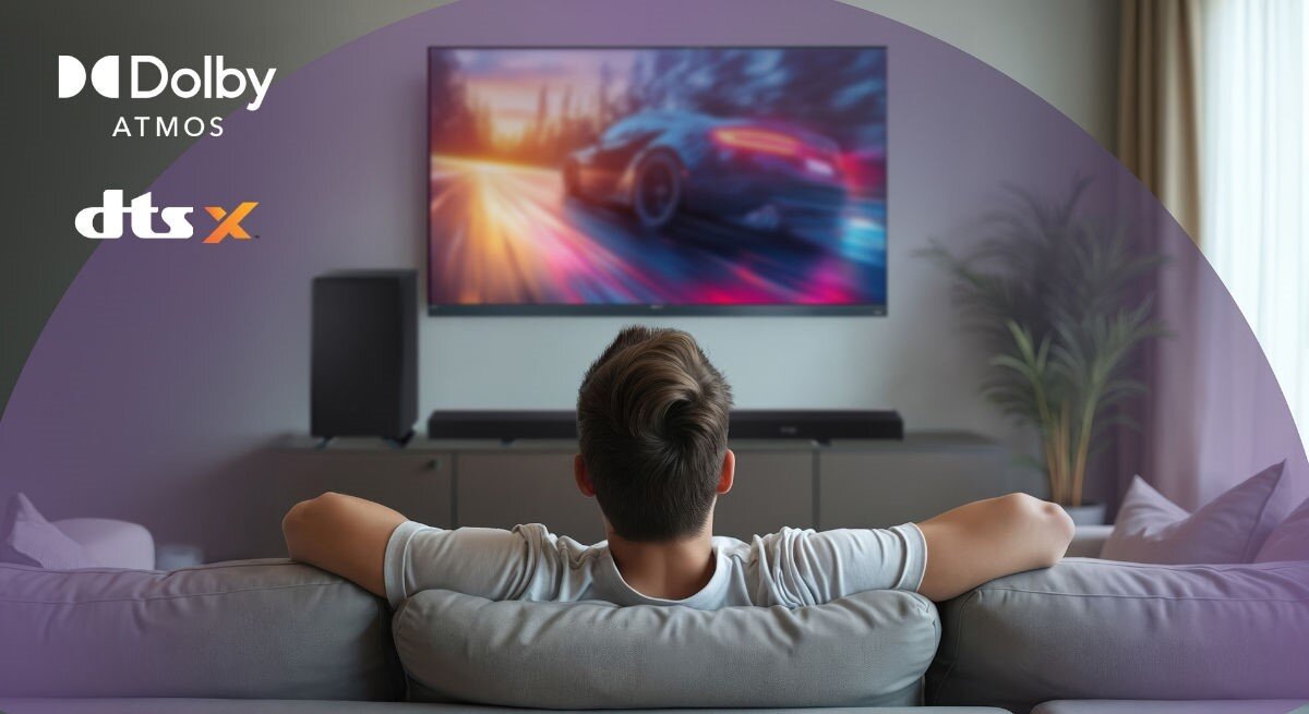 Osoba siedzi na kanapie przed telewizorem, na którym wyświetlany jest dynamiczny obraz samochodu. W lewym górnym rogu widoczne są loga \'Dolby Atmos\' i \'dts X\'.