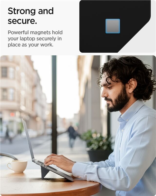 Mężczyzna pracuje na laptopie ustawionym na czarnym stojaku na stole w kawiarni. Tekst na obrazie: \'Strong and secure. Powerful magnets hold your laptop securely in place as you work.\'