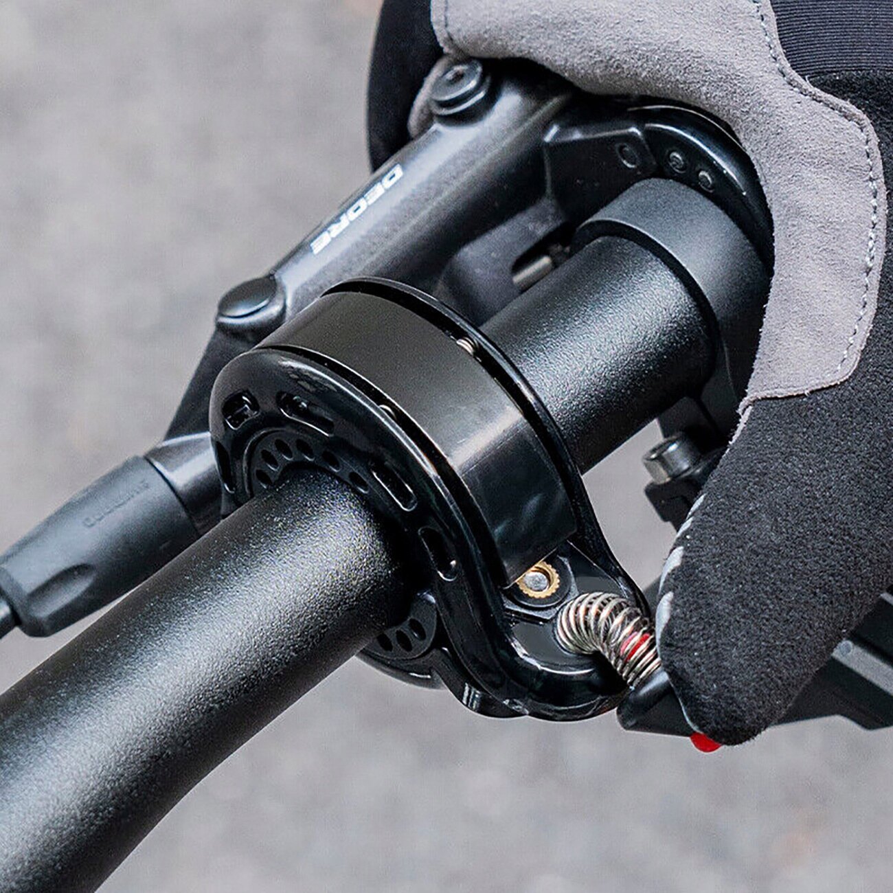 Czarny dzwonek rowerowy zamontowany na kierownicy, z widocznym mechanizmem sprężynowym. Obok znajduje się ręka w rękawiczce trzymająca kierownicę.