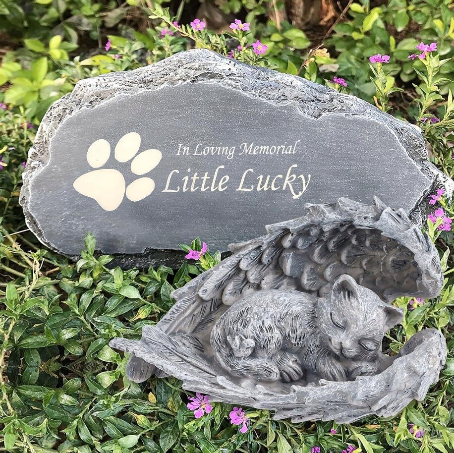 Na obrazie widoczna jest kamienna tablica z napisem \'In Loving Memorial Little Lucky\' oraz figurka śpiącego kota otoczonego skrzydłami anioła, umieszczona na tle zieleni i kwiatów.