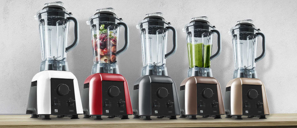 Na drewnianej powierzchni ustawiono pięć blenderów kielichowych w różnych kolorach: biały, czerwony, grafitowy, brązowy i złoty. Każdy blender ma przezroczysty kielich z pokrywką i uchwytem, a w dwóch z nich znajdują się owoce i zielony płyn.