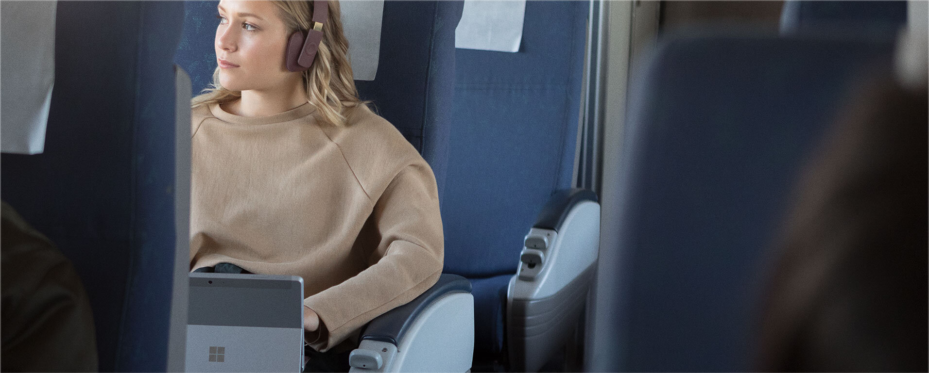 Osoba siedzi w pociągu, mając na sobie słuchawki i trzymając laptop z widocznym logo Microsoft. Siedzenia są pokryte ciemnym materiałem, a na zagłówkach znajdują się białe pokrowce.