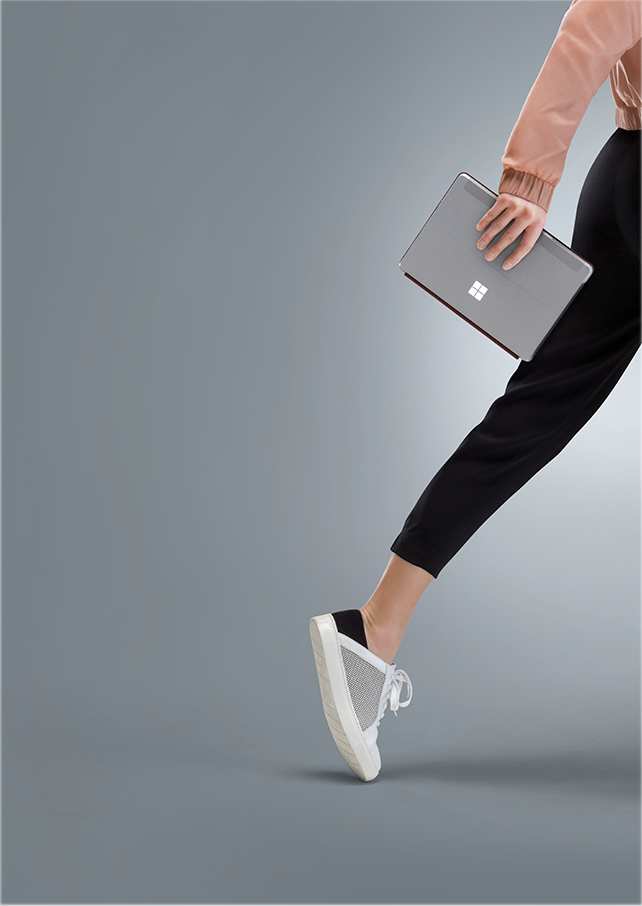 Osoba trzymająca srebrny laptop z widocznym logo w kształcie czterech kwadratów. Osoba ubrana jest w czarne spodnie i białe buty sportowe.