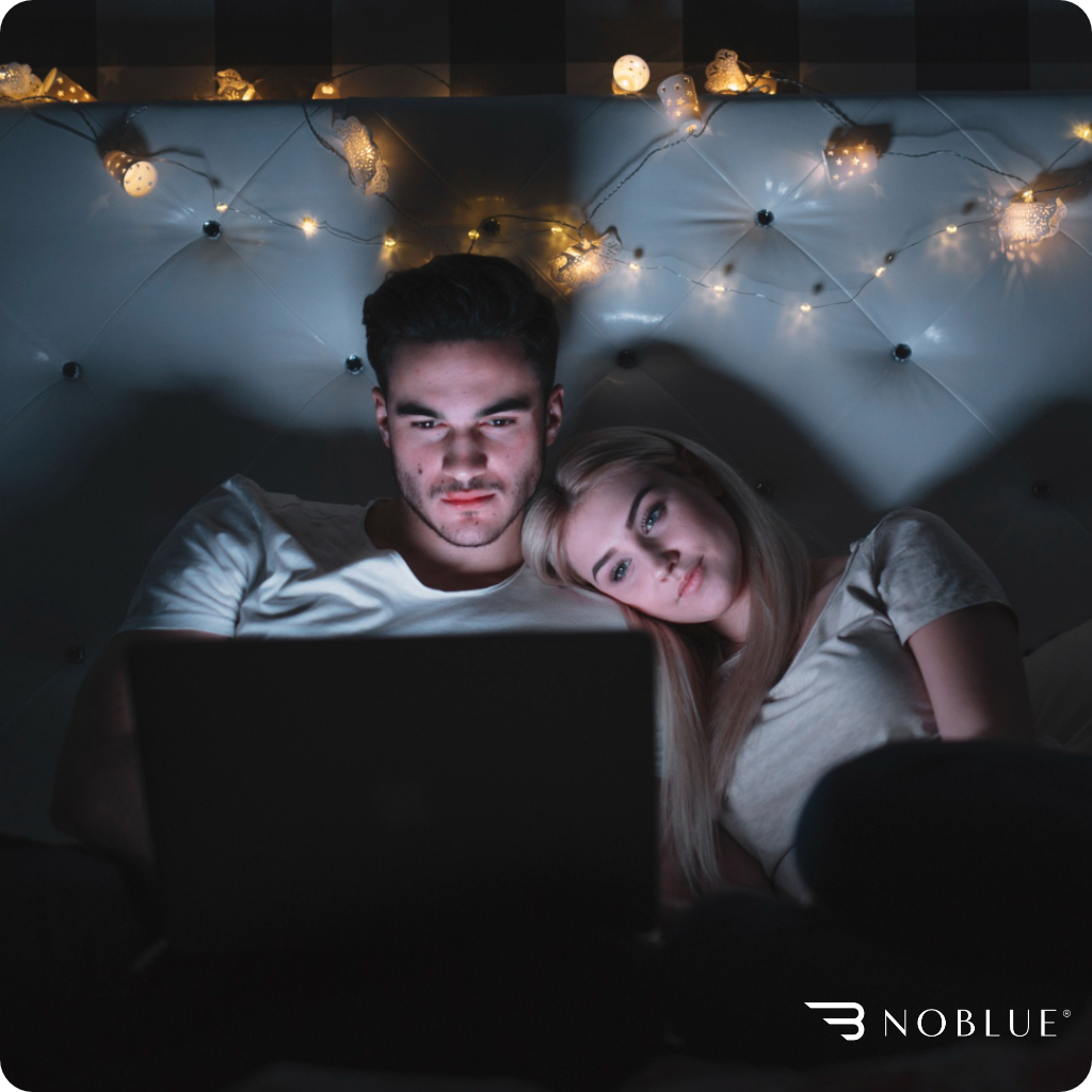 Dwie osoby siedzą na łóżku, patrząc na ekran laptopa. W tle widać światełka dekoracyjne, a w prawym dolnym rogu znajduje się logo \'NOBLUE\'.