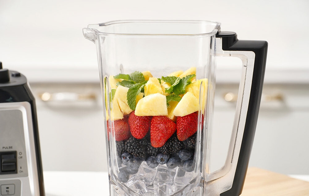 Na obrazie widoczny jest blender kielichowy z przezroczystym pojemnikiem, w którym znajdują się kawałki ananasa, truskawki, jeżyny, borówki i liście mięty. Obok blendera widoczny jest fragment panelu sterowania z przyciskiem \'PULSE\'.