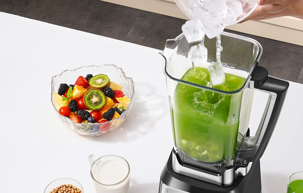 Na blacie stoi blender kielichowy z zielonym płynem, do którego wsypywany jest lód. Obok znajduje się miska z różnorodnymi owocami oraz szklanka z mlekiem.