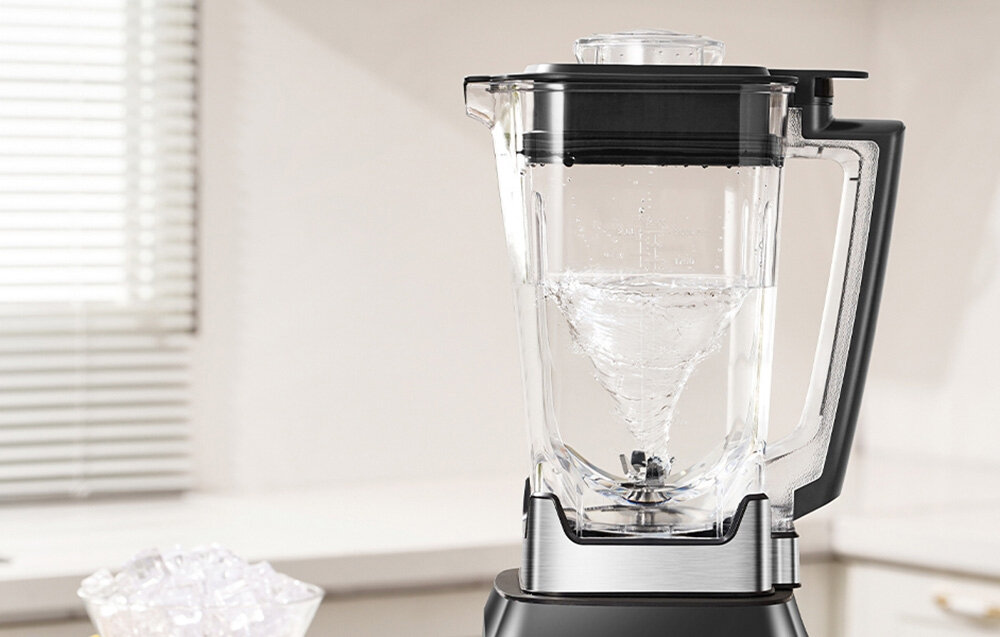Na obrazie widoczny jest blender kielichowy z przezroczystym pojemnikiem, w którym znajduje się wirująca ciecz. W tle widać rozmytą kuchnię z oknem i żaluzjami.