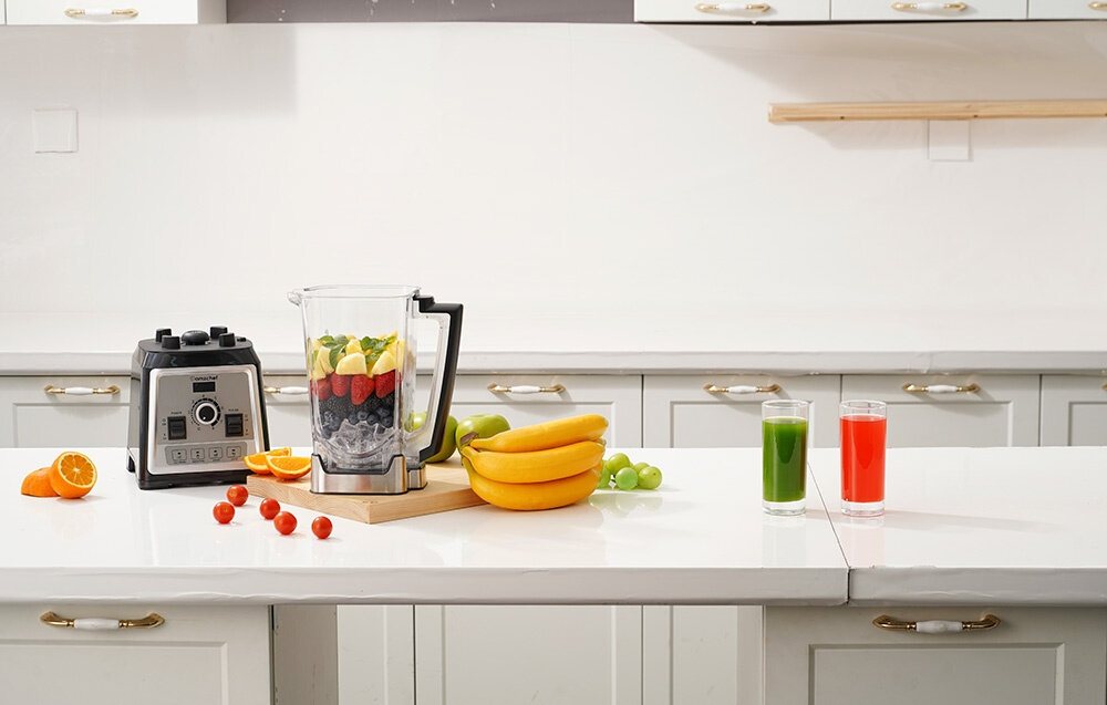 Na blacie kuchennym stoi blender kielichowy z owocami w środku, obok niego leżą banany, pomidorki koktajlowe i połówka pomarańczy. Obok blendera znajdują się dwa szklanki z zielonym i czerwonym napojem.