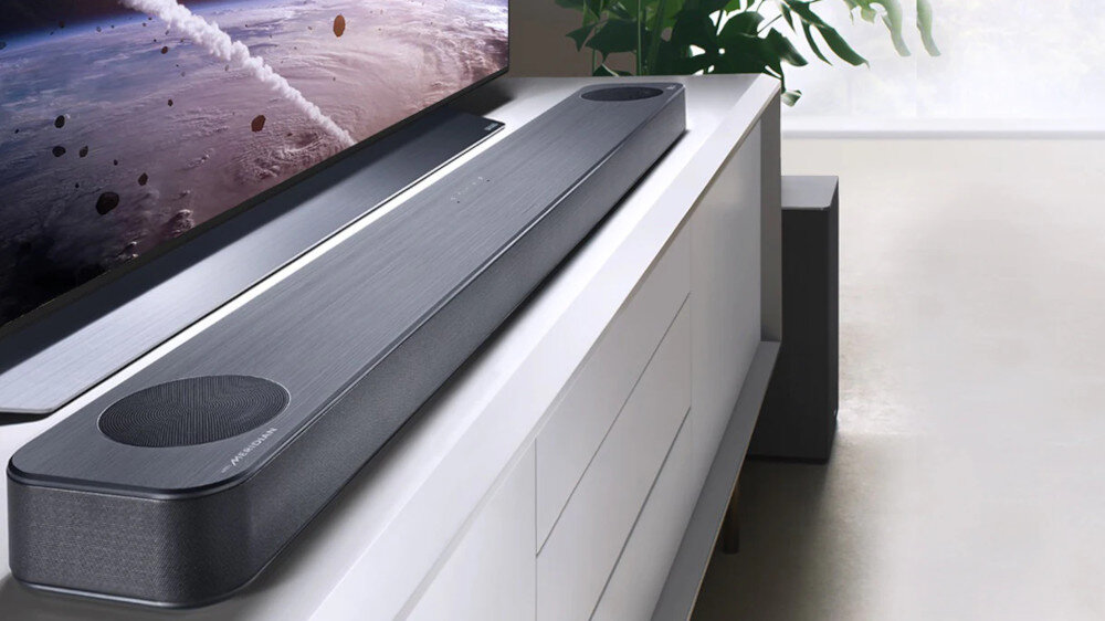 Na białej szafce znajduje się czarny soundbar o nowoczesnym designie. W tle widoczny jest fragment telewizora z obrazem kosmosu oraz rośliny doniczkowe.