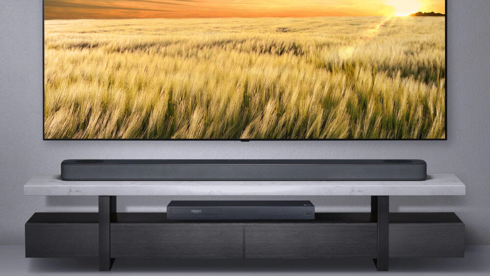 Na obrazie widać czarny soundbar umieszczony na białej półce, poniżej dużego telewizora wyświetlającego pole zboża o zachodzie słońca. Poniżej soundbara znajduje się ciemna szafka RTV.