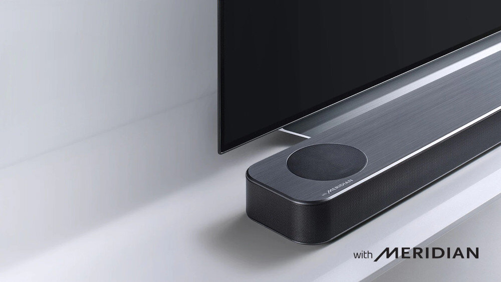 Czarny soundbar z widocznym napisem \'with MERIDIAN\' umieszczony pod telewizorem. Na górnej powierzchni soundbara znajduje się okrągły głośnik.