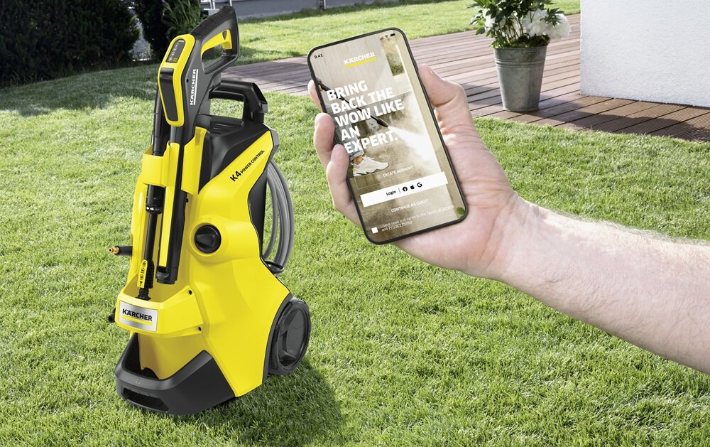 Na trawniku stoi żółta myjka ciśnieniowa z widocznym logo KARCHER. Obok osoba trzyma smartfon z tekstem: \'BRING BACK THE WOW LIKE AN EXPERT\'.