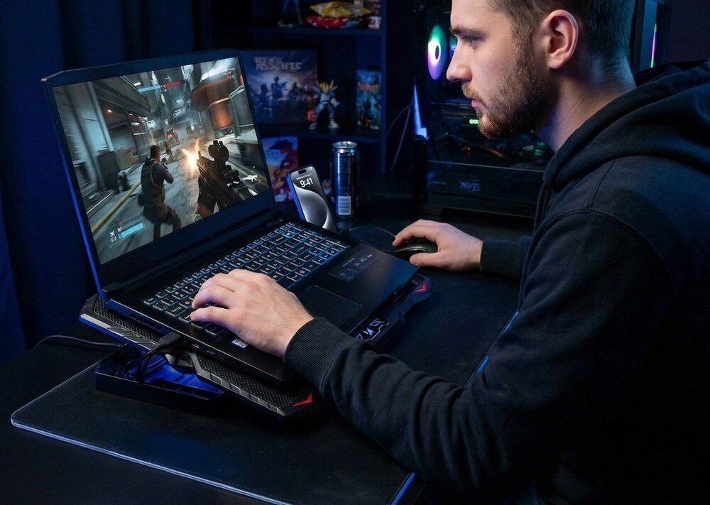 Osoba siedzi przy biurku, używając laptopa umieszczonego na czarnej podstawce chłodzącej z podświetleniem RGB. Na ekranie laptopa widoczna jest gra wideo z postaciami w akcji.