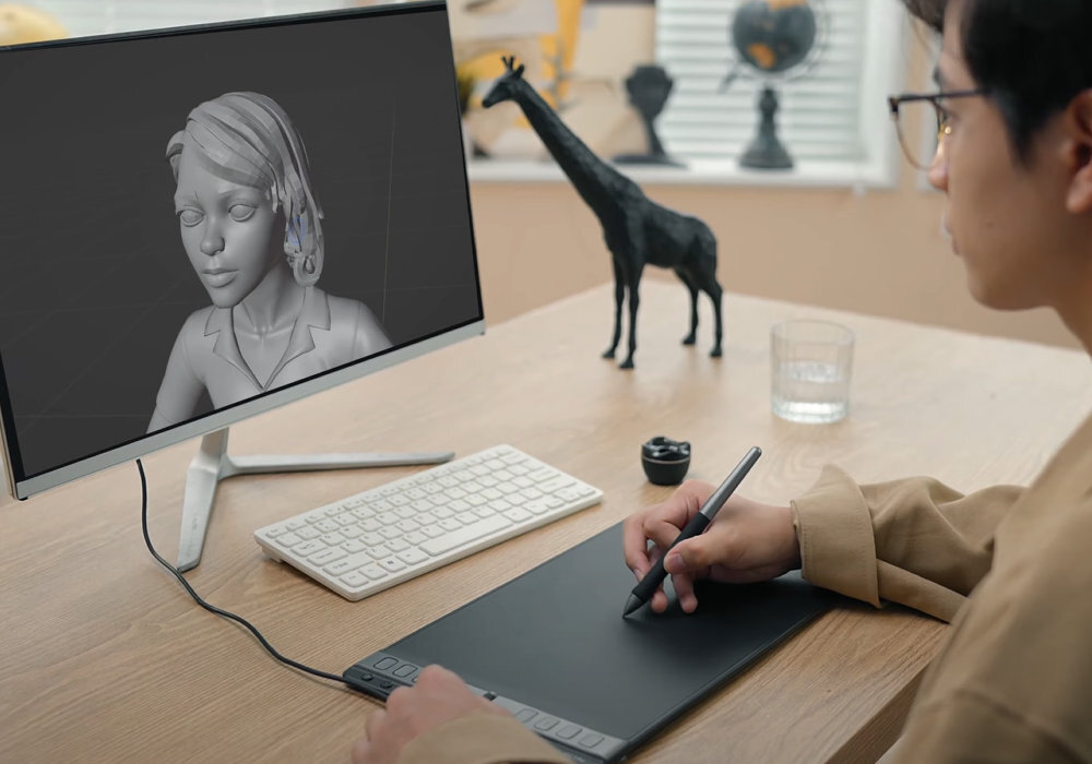 Osoba używa czarnego tabletu graficznego z rysikiem, patrząc na monitor z wyświetlonym modelem 3D postaci. Na biurku znajduje się klawiatura, mysz, szklanka z wodą i figurka żyrafy.