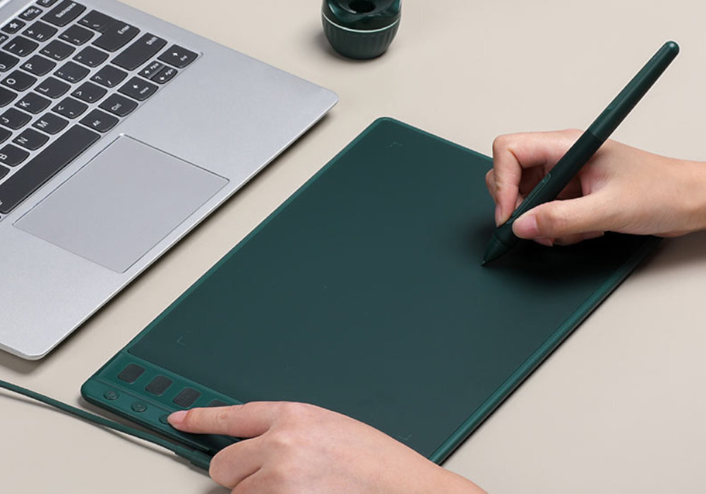 Osoba używa ciemnozielonego tabletu graficznego z rysikiem, podłączonego do laptopa. Na tablecie widoczne są przyciski funkcyjne.