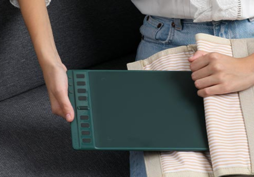 Osoba trzyma czarny tablet graficzny z kilkoma przyciskami po jednej stronie, siedząc na szarej kanapie. Obok znajduje się materiał w paski.