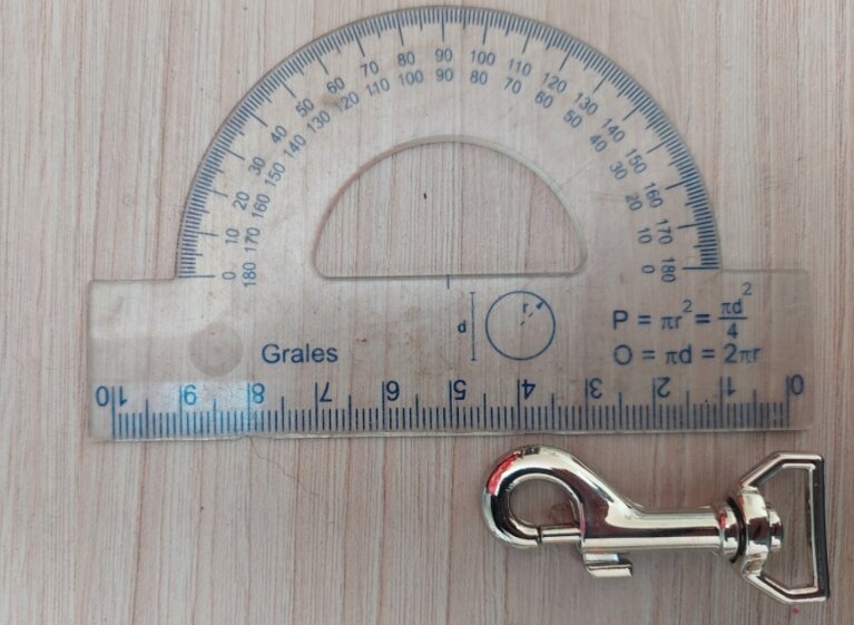 Na drewnianym tle leży przezroczysty kątomierz z nadrukowanymi miarami i wzorami matematycznymi oraz metalowy karabińczyk. Na kątomierzu widoczny jest napis \'Grales\'.