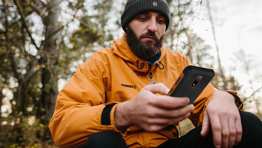 Osoba w pomarańczowej kurtce z napisem \'HAMMER\' trzyma smartfon w ręku. W tle widoczne są drzewa.
