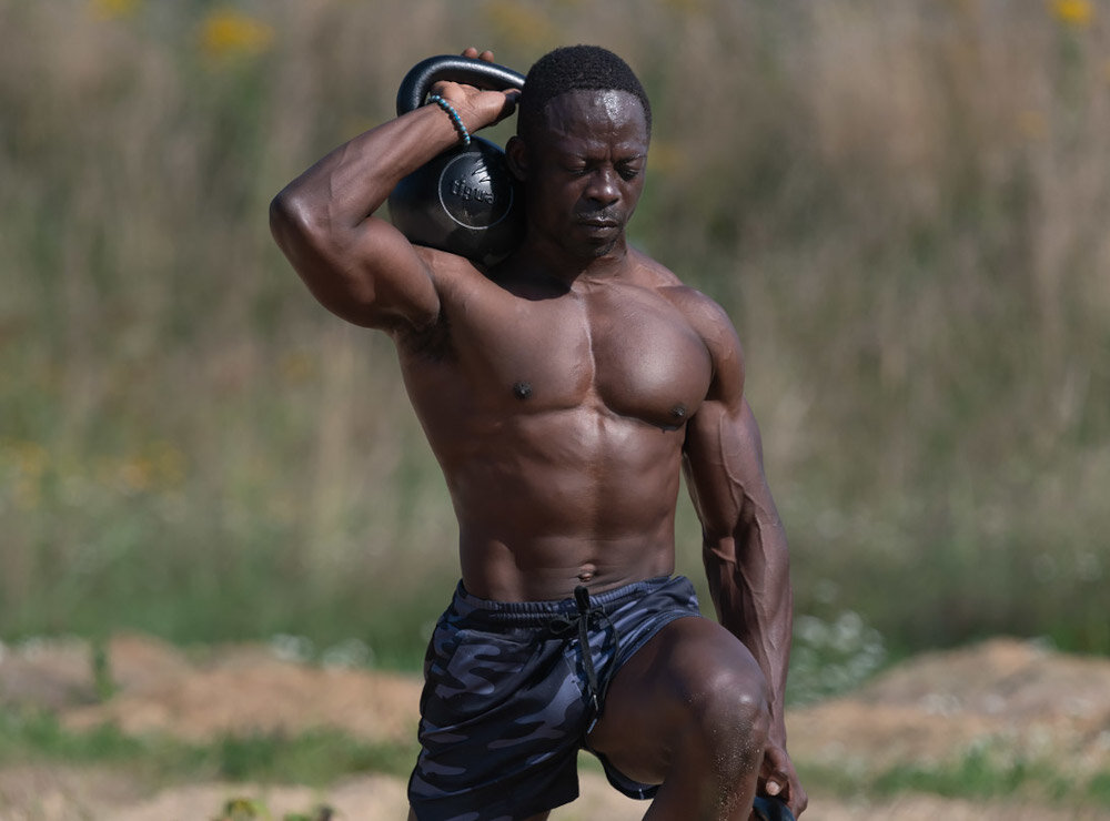 Osoba ćwiczy na świeżym powietrzu, trzymając kettlebell na ramieniu. W tle widać rozmytą roślinność.