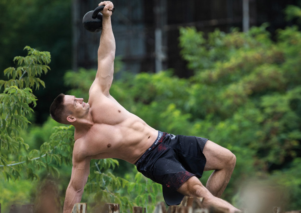 Osoba ćwiczy na świeżym powietrzu, trzymając kettlebell w wyciągniętej ręce. W tle widoczna jest zielona roślinność.