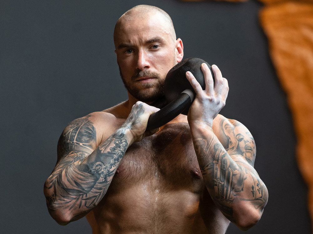 Osoba trzyma kettlebell przy ramieniu, w tle widoczna jest ściana w ciemnym kolorze. Osoba ma tatuaże na ramionach.