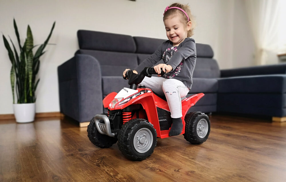 Dziecko siedzi na czerwonym quadzie elektrycznym w pomieszczeniu z drewnianą podłogą i szarą sofą w tle. Obok znajduje się roślina w białej doniczce.