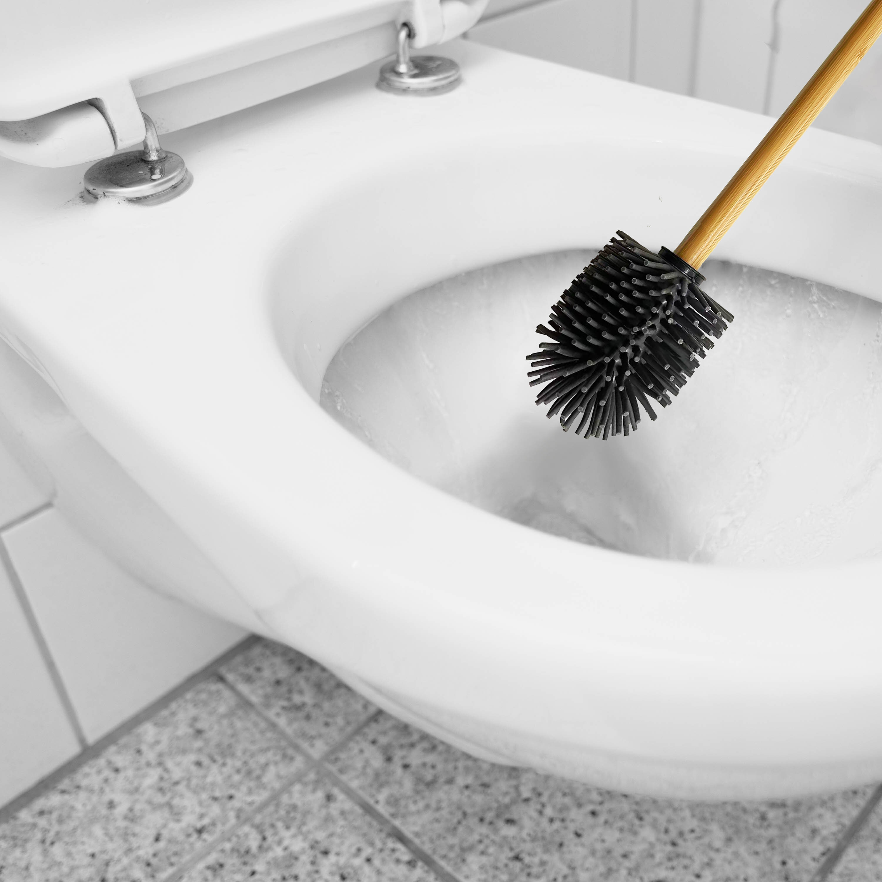 Na zdjęciu widać szczotkę do WC z silikonową główką i drewnianym uchwytem, umieszczoną wewnątrz muszli klozetowej. Podłoga jest wyłożona płytkami.