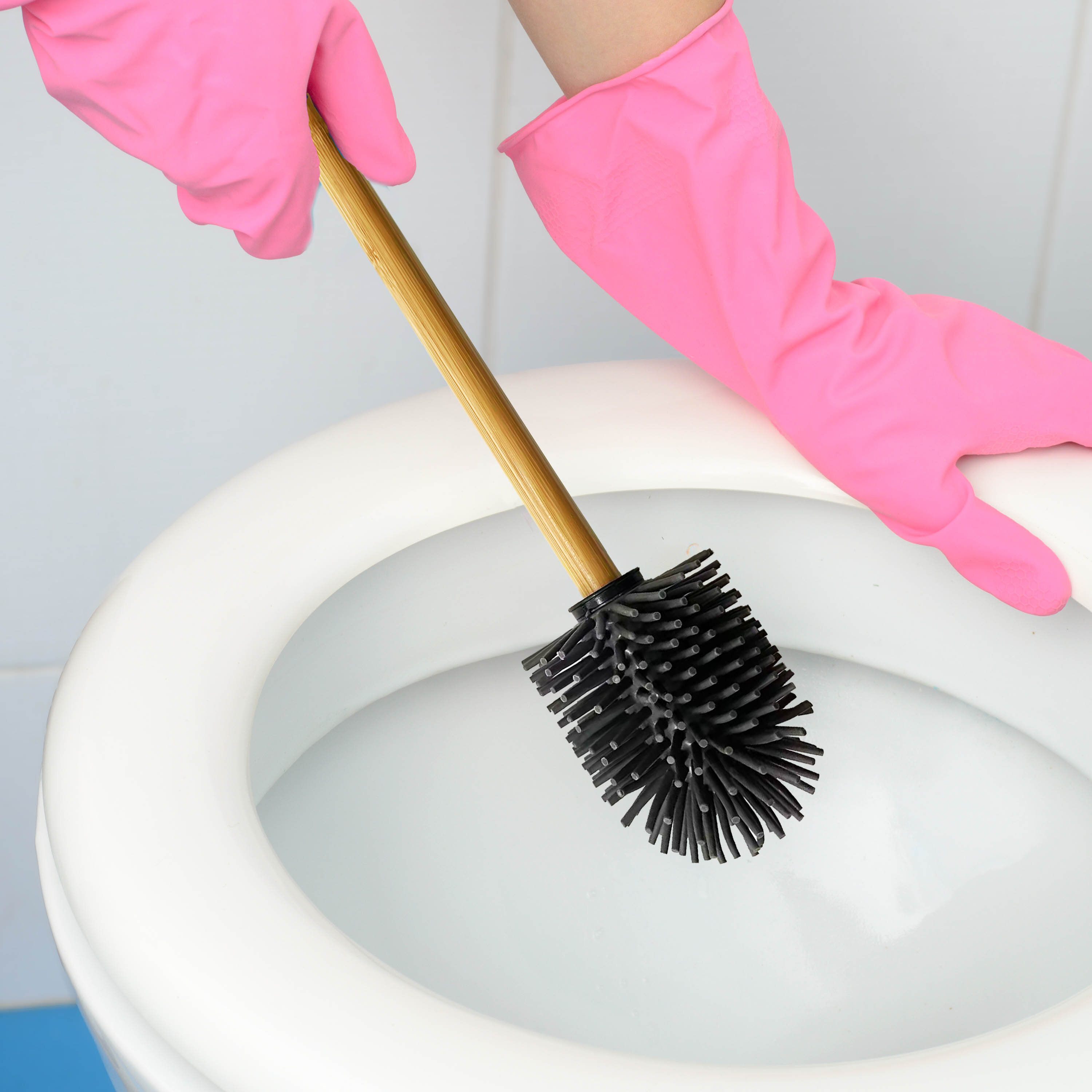 Osoba w różowych rękawiczkach trzyma szczotkę do WC z silikonową główką i drewnianym uchwytem nad muszlą klozetową.