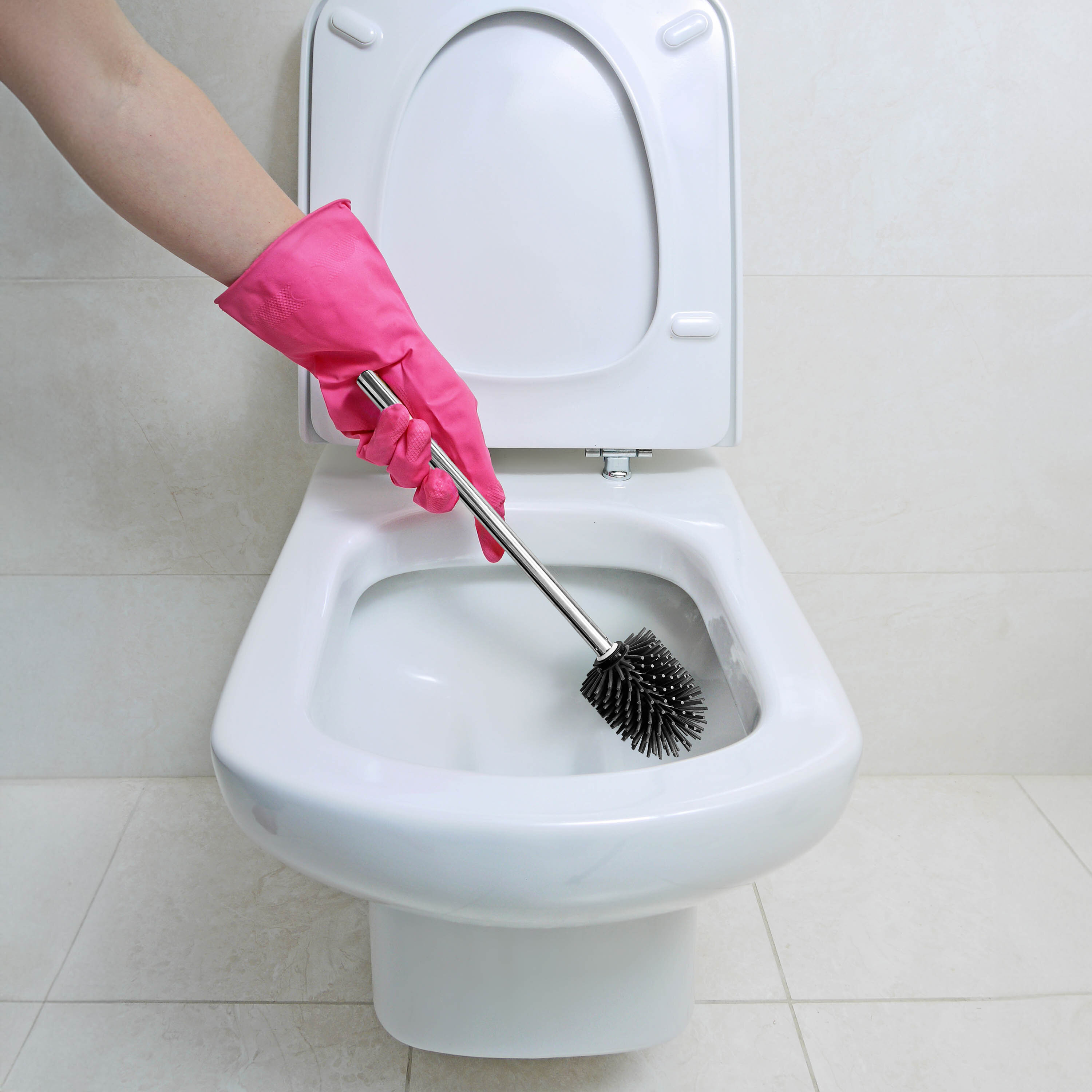 Osoba w różowej rękawicy trzyma szczotkę do WC z czarną silikonową główką nad muszlą klozetową. Tło stanowi jasna, kafelkowa ściana.