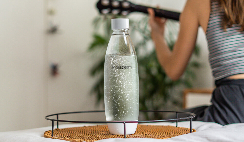 Na zdjęciu znajduje się przezroczysta butelka z napisem \'sodastream\', wypełniona gazowaną wodą, ustawiona na okrągłej podstawce. W tle widoczna jest osoba grająca na gitarze oraz roślina.