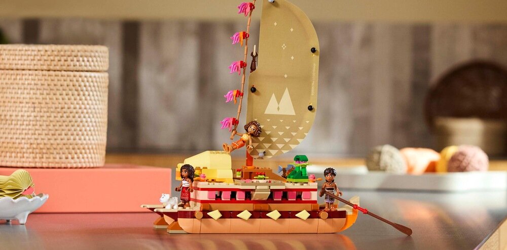 Zestaw LEGO przedstawiający kolorową łódź z żaglem, na której znajdują się dwie figurki postaci. W tle widoczne są rozmyte elementy dekoracyjne.