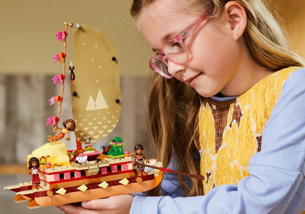 Dziecko trzyma model statku z klocków LEGO z figurkami i żaglem ozdobionym wzorami. Na statku znajdują się różne detale, w tym figurki postaci i ozdoby.