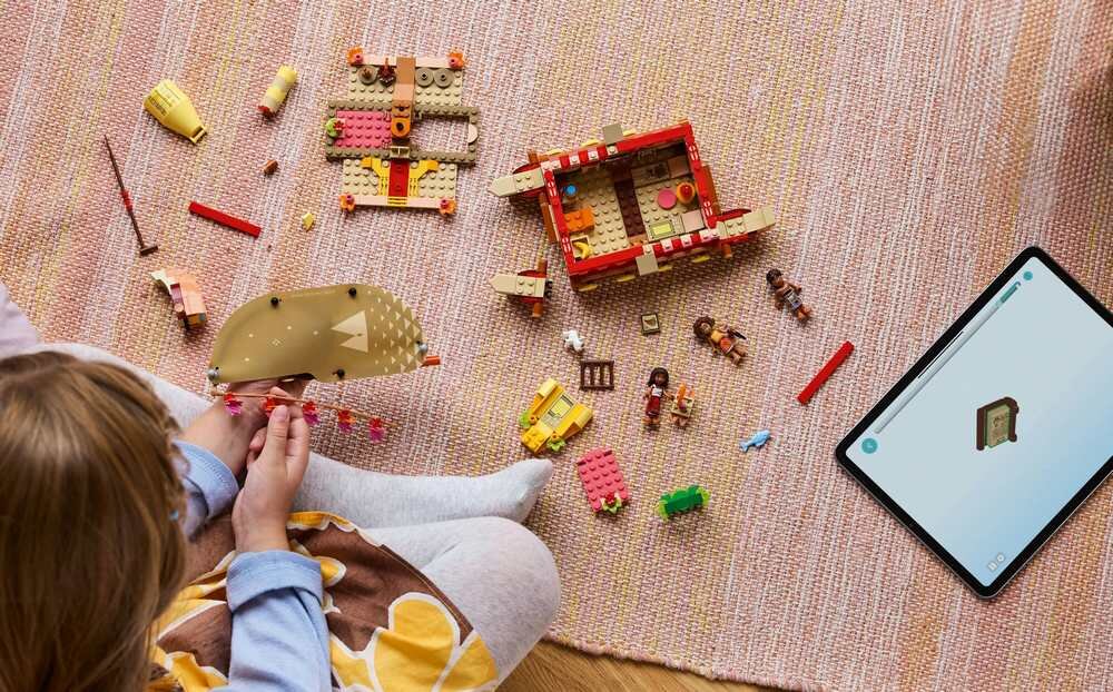 Na dywanie leżą elementy zestawu LEGO, w tym figurki i części konstrukcyjne. Obok znajduje się tablet z wyświetlonym obrazem klocka LEGO.
