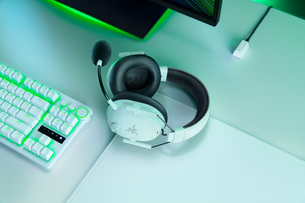Na biurku znajdują się białe słuchawki z mikrofonem oraz klawiatura z podświetleniem. Na słuchawkach widoczne jest logo Razer.