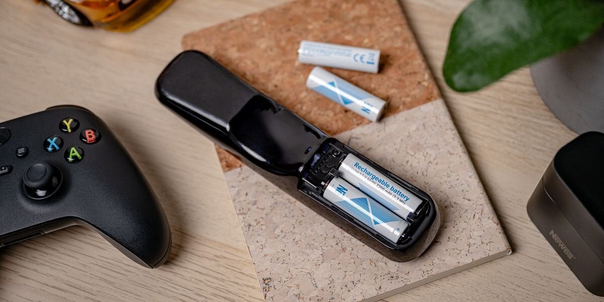 Na drewnianym stole leży czarny kontroler do gier oraz otwarty pilot z dwoma widocznymi akumulatorami z napisem \'Rechargeable battery\'.