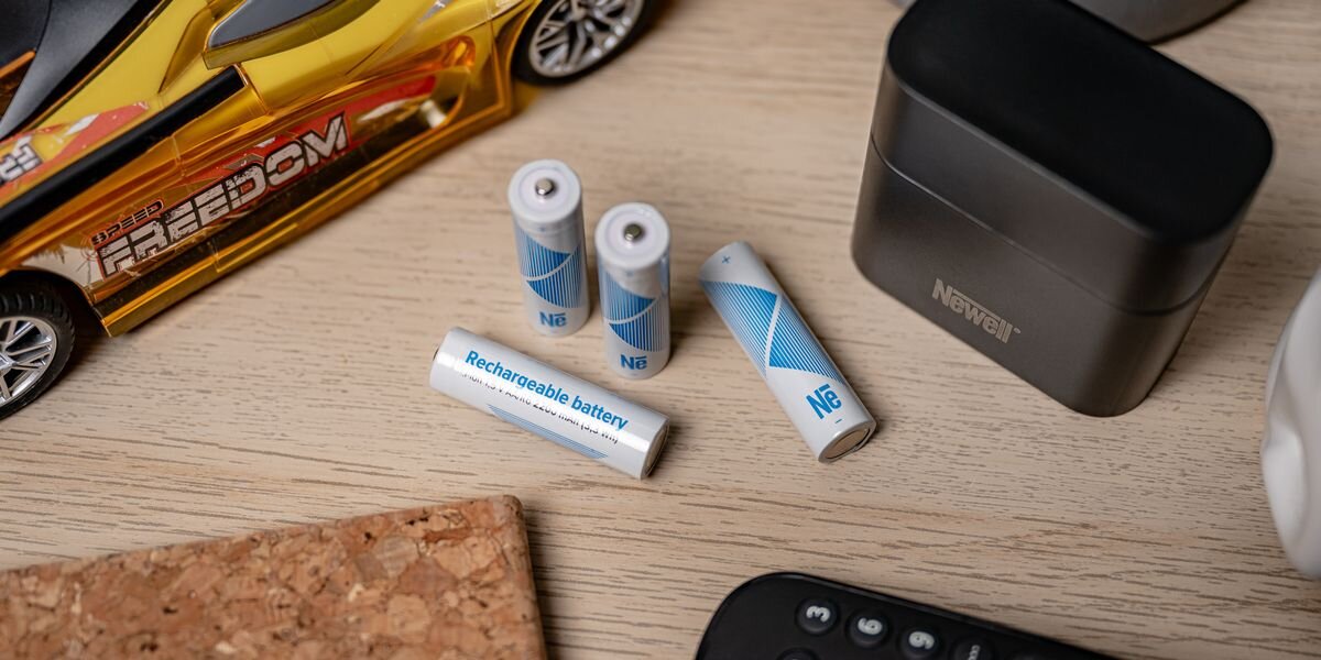 Na drewnianym blacie leżą cztery akumulatory z napisem \'Rechargeable battery\', obok znajduje się model samochodu i czarne etui z logo \'Newell\'.