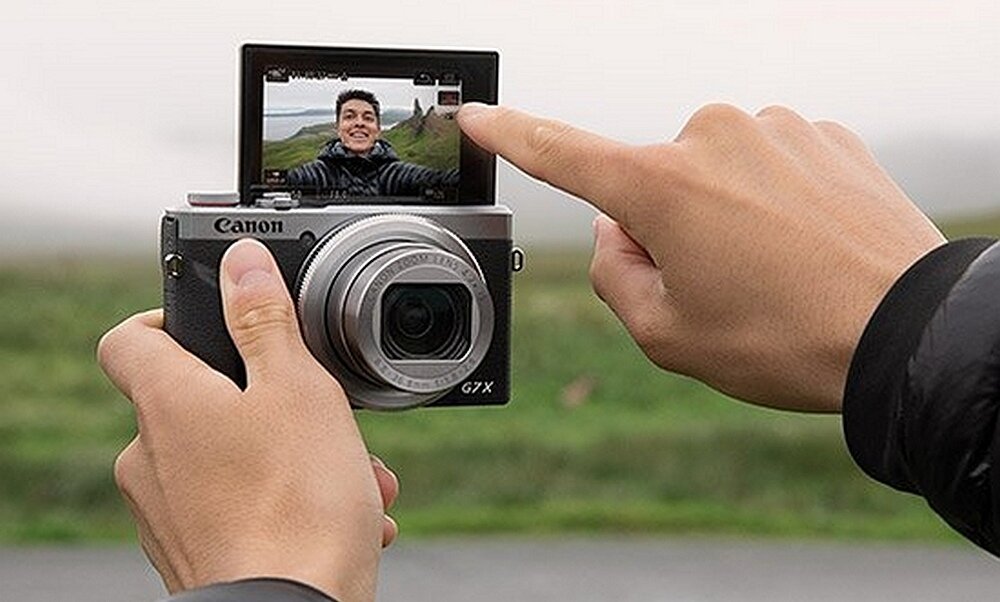 Osoba trzyma aparat fotograficzny z odchylanym ekranem, na którym widoczny jest obraz twarzy. Na aparacie widnieje napis \'Canon G7X\'.
