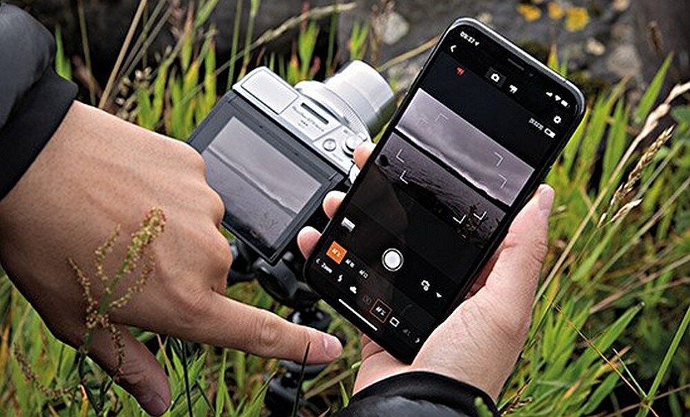 Osoba trzyma smartfon z aplikacją do fotografowania, obok znajduje się aparat fotograficzny na statywie. W tle widoczne są trawy i niebo.