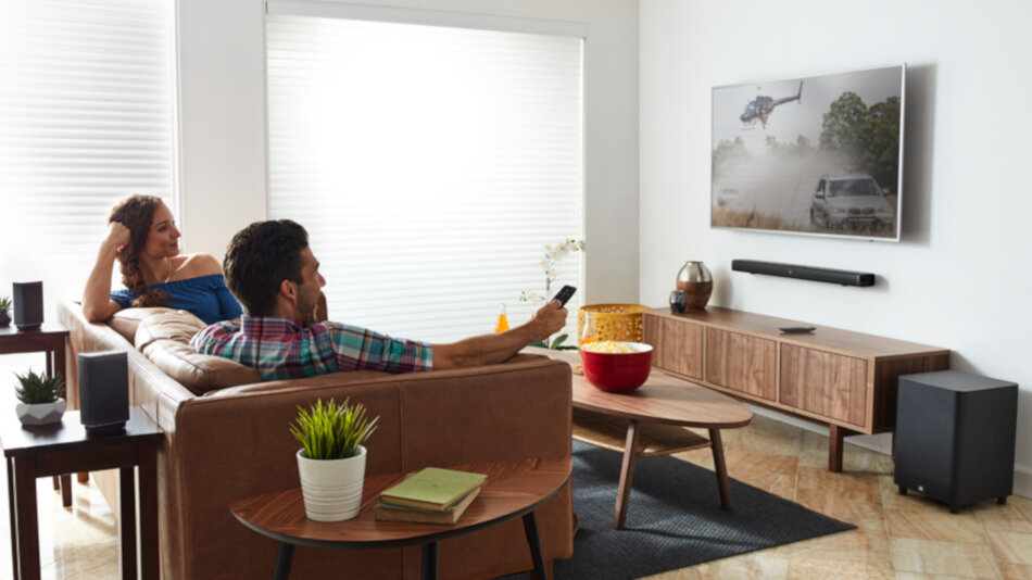 W salonie znajduje się telewizor zamontowany na ścianie, pod którym umieszczony jest czarny soundbar. Na kanapie siedzą dwie osoby, a na stoliku przed nimi stoi miska z przekąskami.