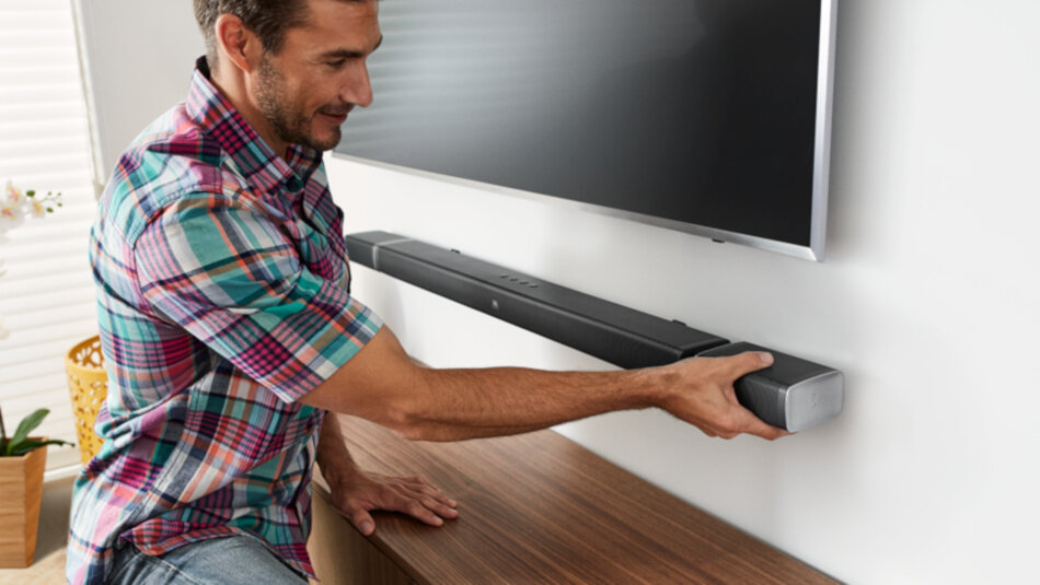 Osoba montuje czarny soundbar pod telewizorem na ścianie. Soundbar ma prostokątny kształt i srebrne końcówki.