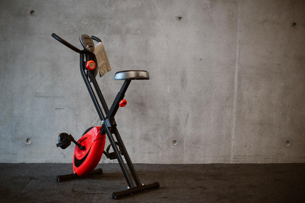 Rower magnetyczny z czarną ramą i czerwonym kołem zamachowym stoi na tle betonowej ściany. Na kierownicy zawieszony jest ręcznik.
