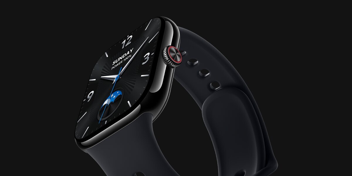 Zbliżenie na czarny smartwatch z zaokrąglonym, błyszczącym ekranem i czarnym paskiem. Na tarczy widoczne są analogowe wskazówki, cyfry 12, 3 i 9, dzień Sunday oraz poziom baterii 100%. Z boku koperty widać pokrętło z czerwonym akcentem i dodatkowy przycisk. Grafika prezentuje wygląd zegarka, jego ekran oraz elementy sterowania.