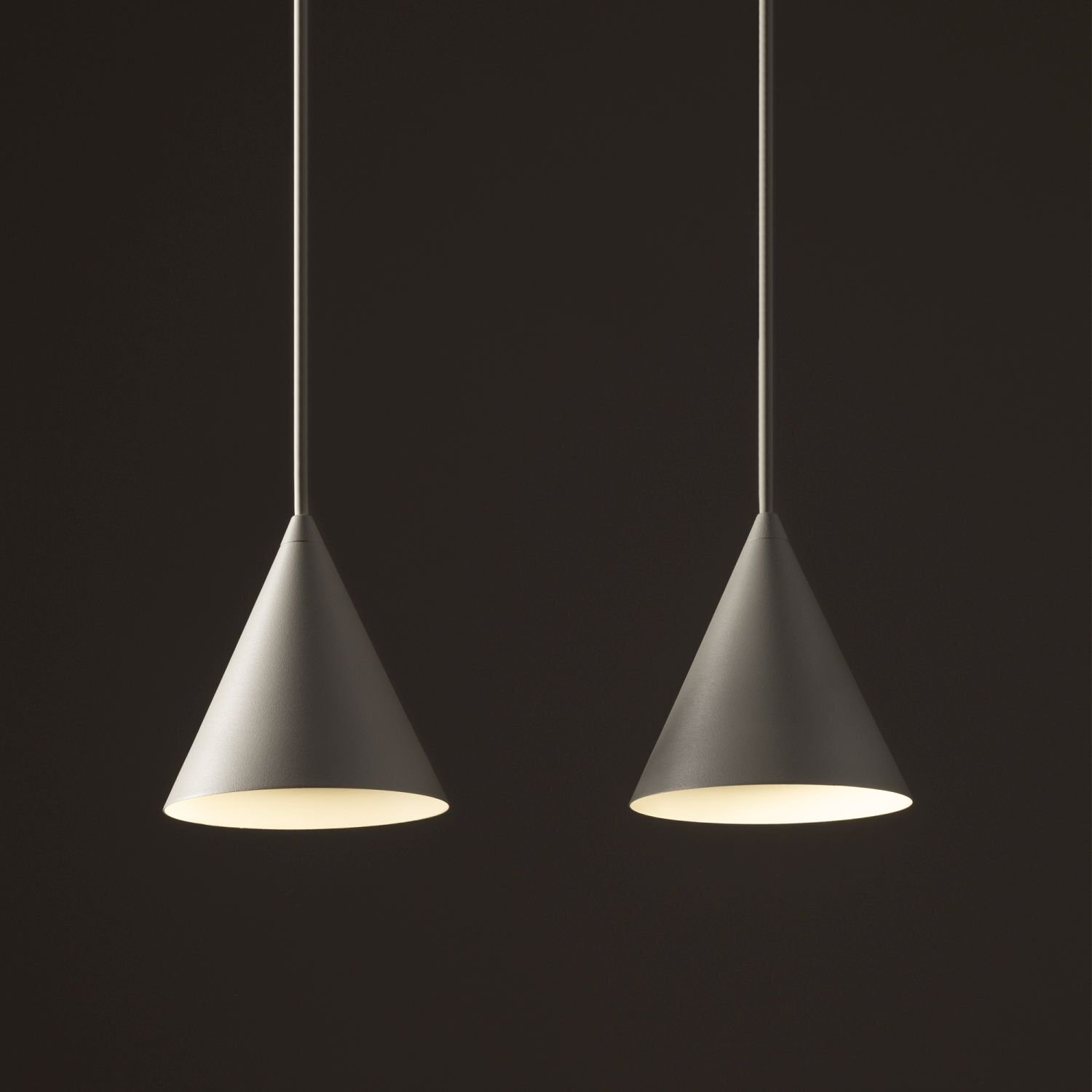 Dwie wiszące lampy w kształcie stożków, wykonane z metalu, emitujące ciepłe światło. Tło jest ciemne, co podkreśla jasność lamp.