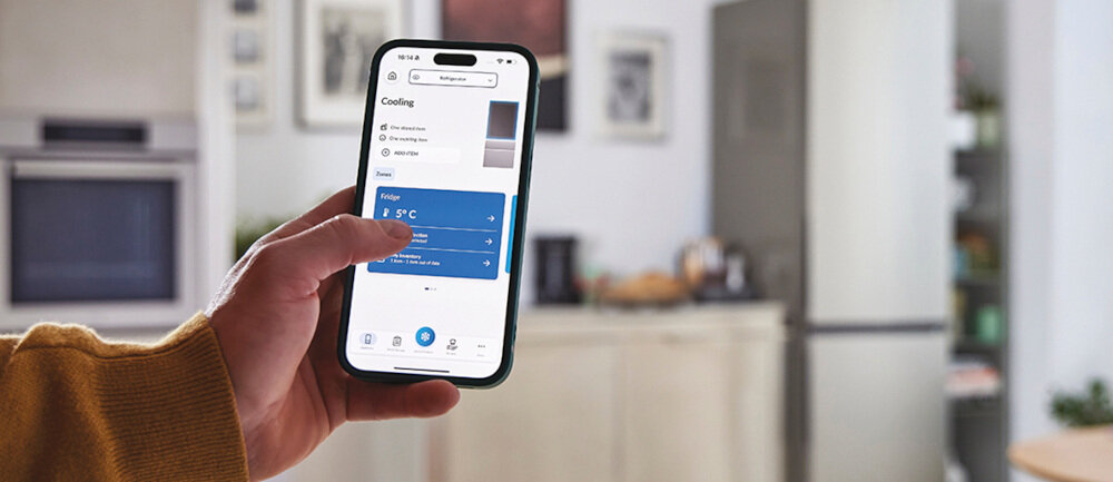 Osoba trzyma smartfon z aplikacją pokazującą ustawienia chłodzenia lodówki. W tle widać nowoczesną kuchnię z lodówką w kolorze inox.