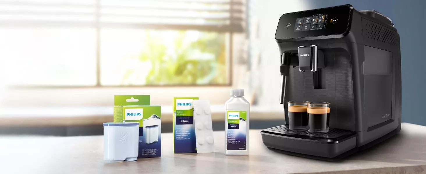 Na blacie znajduje się czarny ekspres do kawy z logo PHILIPS, obok którego stoją dwa kubki z kawą. Obok ekspresu widoczne są opakowania z akcesoriami i środkami czyszczącymi marki PHILIPS.