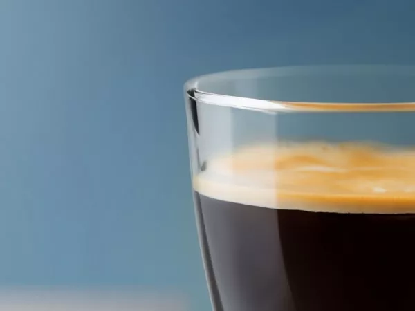 Szklanka z ciemnym napojem, prawdopodobnie kawą, z jasną pianką na górze, na tle niebieskiej ściany.